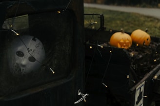 A spooky scene with a white mask with dark eye sockets visible through a dirty window. There are Halloween pumpkins with carved faces on the back of an old vehicle, surrounded by string lights. The atmosphere is eerie and unsettling, enhanced by the dim lighting and weathered textures of the truck.