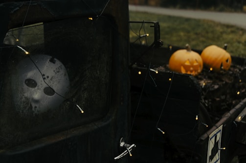 A spooky scene with a white mask with dark eye sockets visible through a dirty window. There are Halloween pumpkins with carved faces on the back of an old vehicle, surrounded by string lights. The atmosphere is eerie and unsettling, enhanced by the dim lighting and weathered textures of the truck.