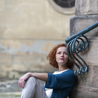 Woman with curly hair leaning against a rustic wall, looking thoughtfully into the distance.