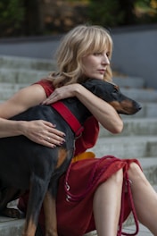 A woman with blonde hair is sitting on outdoor steps and embracing a black Doberman with a red collar. She is wearing a red dress and appears to be deep in thought.