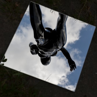 A reflective surface on the ground displays the image of a sky with scattered clouds. Within the reflection, a dark, abstract human figure appears in a dynamic pose, creating a surreal and artistic composition.