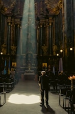 A dimly lit church interior with ornate decorations and high ceilings. Sunlight pours in through a window, illuminating an elderly man standing near the center of the aisle, with rows of empty chairs on either side. A few other figures are seen in the background, and religious icons and flags flank the altar.