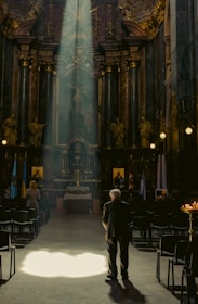 A dimly lit church interior with ornate decorations and high ceilings. Sunlight pours in through a window, illuminating an elderly man standing near the center of the aisle, with rows of empty chairs on either side. A few other figures are seen in the background, and religious icons and flags flank the altar.