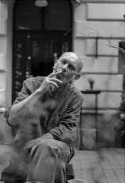 A contemplative man smoking a cigarette sits outdoors, with a blurred background of a menu board and a café setting. The image is in black and white, capturing a candid and pensive moment.