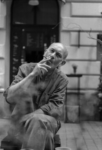 A contemplative man smoking a cigarette sits outdoors, with a blurred background of a menu board and a café setting. The image is in black and white, capturing a candid and pensive moment.