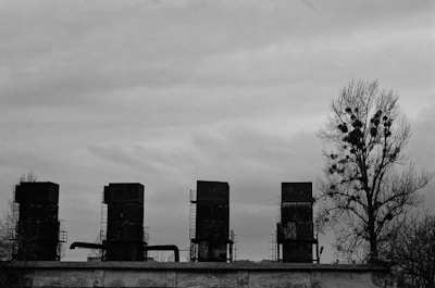 Four large, rectangular industrial chimneys stand on a rooftop. They are tall and old, with metal scaffolding and pipes attached to them. A lone tree with sparse branches and visible nests is situated to the right of the chimneys. The sky is overcast with a blanket of clouds, giving the overall scene a bleak and desolate appearance.