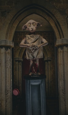 A statue of a small, elf-like creature stands on a pedestal within an archway. The creature has large eyes, long ears, and wears a simple cloth garment. The backdrop is a stone wall that gives a medieval look.