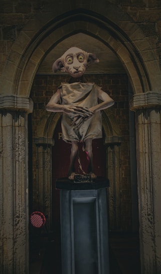 A statue of a small, elf-like creature stands on a pedestal within an archway. The creature has large eyes, long ears, and wears a simple cloth garment. The backdrop is a stone wall that gives a medieval look.