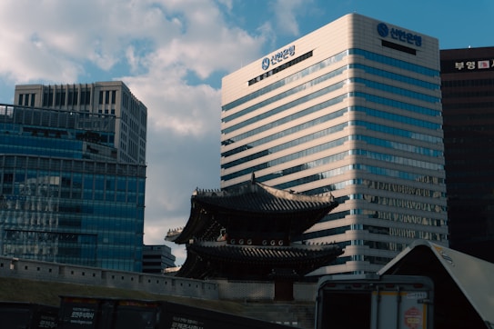 A modern cityscape featuring high-rise buildings with mirrored surfaces and traditional architecture in the form of a historic Korean gate in the middle ground. The sky is partly cloudy, and there is a blend of old and new structures. In the foreground, a section of a parked truck or trailer is visible.