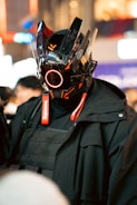 A person is wearing a futuristic helmet featuring sharp angles, glossy black surfaces, and vibrant orange accents. The helmet has a circular red light in the center, and technical details suggest a sci-fi or robotic theme. The individual is dressed in dark clothing with a heavy, tactical appearance.