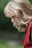 A woman with blonde hair is captured in profile against a blurred green background. She wears a red garment, and her hair falls in loose waves over her shoulders, partially obscuring her face as she gazes downward.
