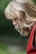 A woman with blonde hair is captured in profile against a blurred green background. She wears a red garment, and her hair falls in loose waves over her shoulders, partially obscuring her face as she gazes downward.