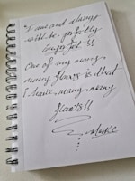 A spiral-bound notebook with handwritten text in dark ink. The writing expresses a message about embracing imperfections and acknowledges having many flaws. The handwriting is cursive and artistic, emphasizing the unique and personal nature of the note.