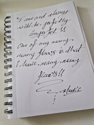 A spiral-bound notebook with handwritten text in dark ink. The writing expresses a message about embracing imperfections and acknowledges having many flaws. The handwriting is cursive and artistic, emphasizing the unique and personal nature of the note.