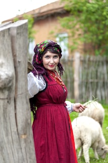 A person dressed in traditional clothing is standing outdoors, leaning against a wooden fence. The outfit features a burgundy dress and a colorful headscarf. In the background, there are sheep grazing in a green, grassy area, and a rustic building is partially visible.