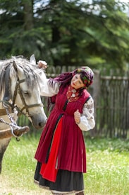 A person dressed in traditional clothing with intricate embroidery and a colorful headscarf stands beside a grey horse. The background features lush green foliage and a wooden fence.