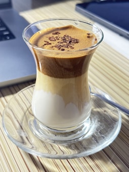 A glass cup filled with a layered coffee beverage, featuring a frothy, whipped coffee layer on top with cocoa or coffee powder garnish, and a milk layer at the bottom. The cup is placed on a clear glass saucer on a striped table surface, with part of a laptop visible in the background.