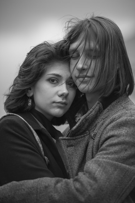 A monochrome photograph of two young individuals in an intimate embrace, with expressions conveying a sense of connection and emotion. The wind gently tussles their hair, adding a dynamic element to the composition. They are dressed in warm clothing, suggesting a cold environment.