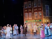 A theatrical scene with actors dressed in period costumes performing in front of a set resembling an old pub called George's Pub. The building facade is detailed with brick and window designs, and several people are gathered, wearing hats and nineteenth-century attire.