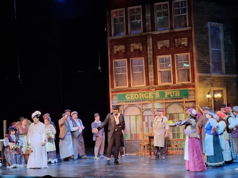 A theatrical scene with actors dressed in period costumes performing in front of a set resembling an old pub called George's Pub. The building facade is detailed with brick and window designs, and several people are gathered, wearing hats and nineteenth-century attire.