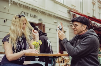 A man wearing glasses and a beret is photographing a woman with long blonde hair sitting across from him at an outdoor café table. The woman, wearing sunglasses and a printed t-shirt, is smiling and holding a small bouquet of yellow flowers placed on the table.