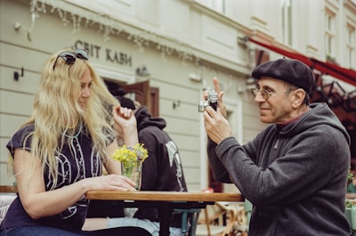 A man wearing glasses and a beret is photographing a woman with long blonde hair sitting across from him at an outdoor café table. The woman, wearing sunglasses and a printed t-shirt, is smiling and holding a small bouquet of yellow flowers placed on the table.