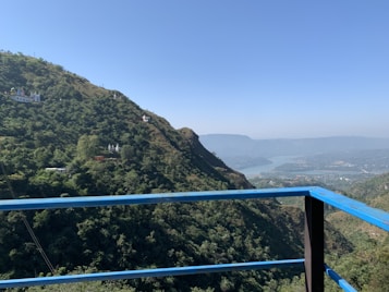 A scenic view of lush green hills with a calm river flowing below, surrounded by distant mountain ranges. A blue railing in the foreground suggests the picture was taken from a balcony or a viewing platform. Sparse structures, possibly temples or small buildings, are visible on the hills.