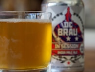 A close-up view of a glass filled with a golden, frothy beverage placed in front of a can with a patriotic-themed design. The can features bold, red and blue typography along with stars, suggestive of an American craft beer.