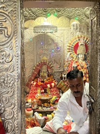 A man dressed in a white shirt is inside a richly decorated shrine adorned with intricate carvings. The shrine contains a statue of a deity, dressed in colorful and ornate attire. Strings of multicolored lights and flowers enhance the decorative scene, and various offerings are placed before the deity, including flowers, incense, and other ritual items.