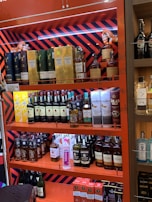 Shelves stocked with various brands of liquor, including bottles of whisky and gin. The display features brands like Dewar's, Monkey Shoulder, Jameson, Chivas, The Glenlivet, and others. The shelves are brightly colored with orange and red patterns.