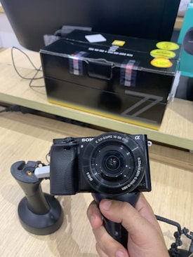 A hand is holding a black digital camera with a zoom lens. The camera is displayed against a retail setting, with a boxed product in the background. The camera lens has specifications written on it. The box in the background is predominantly black with some yellow and red markings.