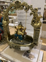 A decorative statue featuring intricately designed elements, showcasing a figure seated on a swing. The swing is adorned with detailed carvings and patterns, with rich use of gold and teal colors. The swing has ornate posts with animal motifs.