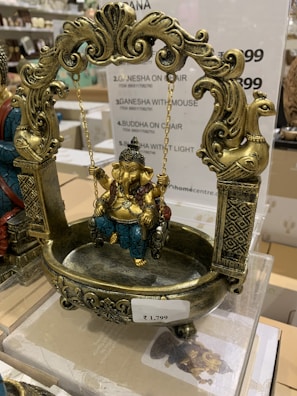 A decorative statue featuring intricately designed elements, showcasing a figure seated on a swing. The swing is adorned with detailed carvings and patterns, with rich use of gold and teal colors. The swing has ornate posts with animal motifs.
