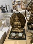 A golden Buddha statue is prominently displayed on a retail shelf surrounded by various other similar spiritual or decorative items. The Buddha statue features intricate detailing and is seated in a meditative pose with both hands in a prayer gesture. The base of the display includes small stones and two candle holders. In the background, there are other statues and items, including another Buddha statue and a Ganesha statue. White boxes with decorative designs are also visible.