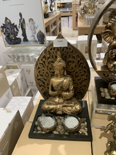 A golden Buddha statue is prominently displayed on a retail shelf surrounded by various other similar spiritual or decorative items. The Buddha statue features intricate detailing and is seated in a meditative pose with both hands in a prayer gesture. The base of the display includes small stones and two candle holders. In the background, there are other statues and items, including another Buddha statue and a Ganesha statue. White boxes with decorative designs are also visible.