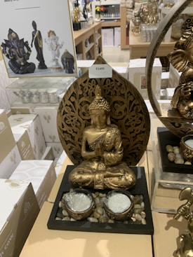 A golden Buddha statue is prominently displayed on a retail shelf surrounded by various other similar spiritual or decorative items. The Buddha statue features intricate detailing and is seated in a meditative pose with both hands in a prayer gesture. The base of the display includes small stones and two candle holders. In the background, there are other statues and items, including another Buddha statue and a Ganesha statue. White boxes with decorative designs are also visible.