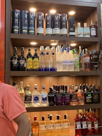 A well-lit wooden liquor shelf filled with a variety of alcoholic beverages. The top shelf displays premium whiskey bottles, including some branded Glenfiddich containers and a bottle of John Barr. The middle shelf features various vodkas, including Ciroc and Grey Goose, and a range of coffee liqueurs. The bottom two shelves contain an assortment of gins, like Bulldog and Jamun, as well as other spirits like Fireball and Toki whisky. A person in a pink shirt stands partially visible on the left side.