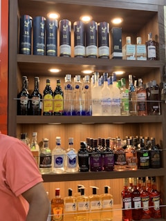 A well-lit wooden liquor shelf filled with a variety of alcoholic beverages. The top shelf displays premium whiskey bottles, including some branded Glenfiddich containers and a bottle of John Barr. The middle shelf features various vodkas, including Ciroc and Grey Goose, and a range of coffee liqueurs. The bottom two shelves contain an assortment of gins, like Bulldog and Jamun, as well as other spirits like Fireball and Toki whisky. A person in a pink shirt stands partially visible on the left side.