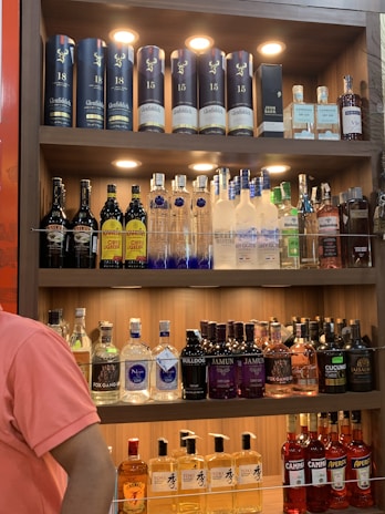 A well-lit wooden liquor shelf filled with a variety of alcoholic beverages. The top shelf displays premium whiskey bottles, including some branded Glenfiddich containers and a bottle of John Barr. The middle shelf features various vodkas, including Ciroc and Grey Goose, and a range of coffee liqueurs. The bottom two shelves contain an assortment of gins, like Bulldog and Jamun, as well as other spirits like Fireball and Toki whisky. A person in a pink shirt stands partially visible on the left side.