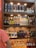 A well-lit wooden liquor shelf filled with a variety of alcoholic beverages. The top shelf displays premium whiskey bottles, including some branded Glenfiddich containers and a bottle of John Barr. The middle shelf features various vodkas, including Ciroc and Grey Goose, and a range of coffee liqueurs. The bottom two shelves contain an assortment of gins, like Bulldog and Jamun, as well as other spirits like Fireball and Toki whisky. A person in a pink shirt stands partially visible on the left side.