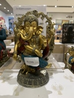 A decorative statue of Lord Ganesha with intricate detailing. The figure is adorned with a headdress and various ornaments, and it is painted in gold with accents of blue and red. It is placed on a decorative base and there is a price tag attached to it.