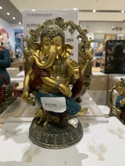 A decorative statue of Lord Ganesha with intricate detailing. The figure is adorned with a headdress and various ornaments, and it is painted in gold with accents of blue and red. It is placed on a decorative base and there is a price tag attached to it.