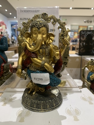 A decorative statue of Lord Ganesha with intricate detailing. The figure is adorned with a headdress and various ornaments, and it is painted in gold with accents of blue and red. It is placed on a decorative base and there is a price tag attached to it.