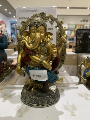 A decorative statue of Lord Ganesha with intricate detailing. The figure is adorned with a headdress and various ornaments, and it is painted in gold with accents of blue and red. It is placed on a decorative base and there is a price tag attached to it.