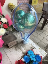 A transparent balloon filled with turquoise balloons is wrapped with blue ribbons, placed next to a bouquet of blue and white flowers. A wooden bench and some potted plants with yellow flowers are nearby. The scene is set on a tiled outdoor area with a plastic chair and a partial view of an advertisement poster in the background.