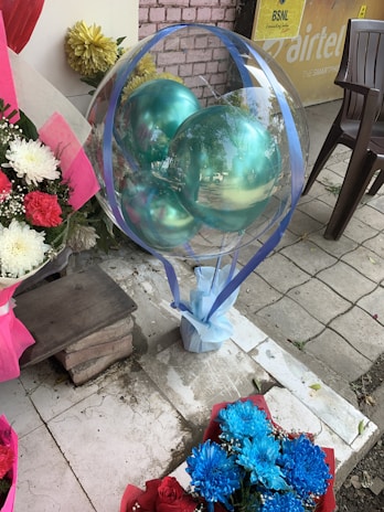 A transparent balloon filled with turquoise balloons is wrapped with blue ribbons, placed next to a bouquet of blue and white flowers. A wooden bench and some potted plants with yellow flowers are nearby. The scene is set on a tiled outdoor area with a plastic chair and a partial view of an advertisement poster in the background.