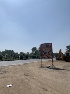 An outdoor scene featuring a large advertising sign for a highway restaurant. The ground appears dry and sandy, with a clear, blue sky above. There is a road adjacent to the sign, bordered by trees and greenery. A construction vehicle, possibly an excavator or backhoe, is partially visible on the right.