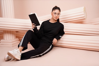 A person wearing a black tracksuit with white stripes is sitting on the floor, leaning against horizontal, off-white classical columns. The individual is holding an open black notebook and is wearing clear glasses. They have their hair tied back in a neat ponytail and are wearing white sneakers.