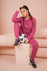 A person with braided hair is sitting on a beige pedestal, dressed in a matching pink tracksuit and black sneakers with purple laces. They are holding a soccer ball with a hand while resting their head on the other hand.