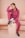 A person with braided hair is sitting on a beige pedestal, dressed in a matching pink tracksuit and black sneakers with purple laces. They are holding a soccer ball with a hand while resting their head on the other hand.